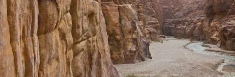 Schlucht mit hohen, braunen Felswänden und einem Kiesbett am Boden und einem kleinen Fluss