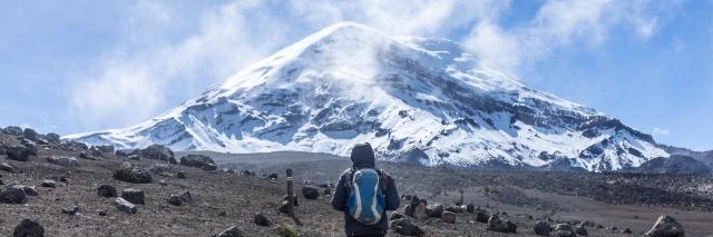 Mensch mit Rucksack steht vor einem schneebedeckten Vulkan in einer grauen Lavalandschaft