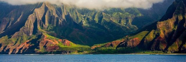 Küstenlandschaft hinter blauem Meer mit hohen, grünbewachsenen Bergen mit Nebel
