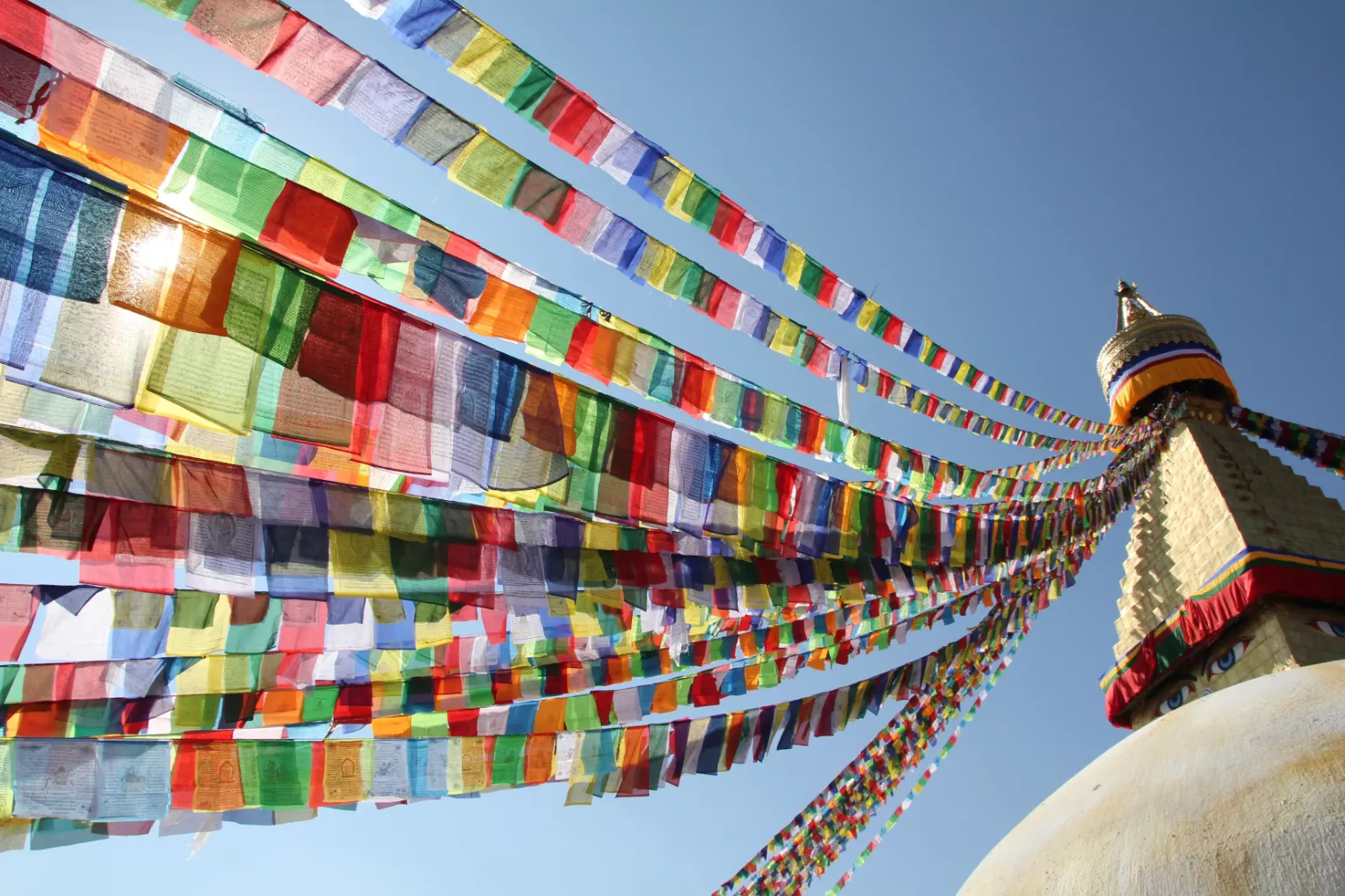 Der Stupa von Bodnath in Nepal ist rechts im Bild zu sehen, von der aus viele bunte Gebetsfahnen zum linken Bildrand aufgehängt sind.
