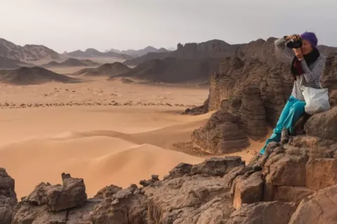 Eine Frau mit Kopftuch sitzt mit einer Kamera rechts vorne im Bild auf einem großen Felsen. Im Hintergrund ist eine Sandwüste zu sehen. Immer wieder ragen große Felsen aus dem Sand hervor. Die Frau blickt durch das Objektiv der Kamera. 