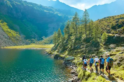 Eine Gruppe von fünf Wanderern geht auf einem schmalen Pfad direkt entlang eines blauen Bergsees. Hinter dem See ist ein hügeliges Tal zu sehen.