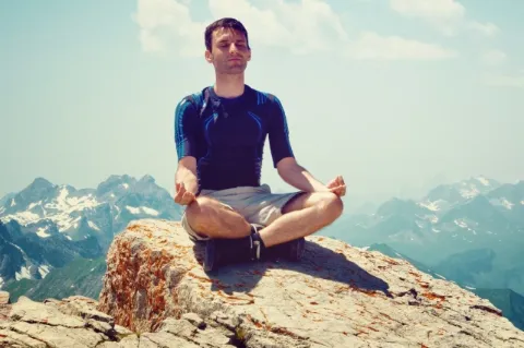 Ein junger Mann sitzt im Schneidersitz auf einem Berggipfel und hält die Hände in Meditationspose. Im Hintergrund ist ein hohes Gebirge mit Schnee zu sehen.