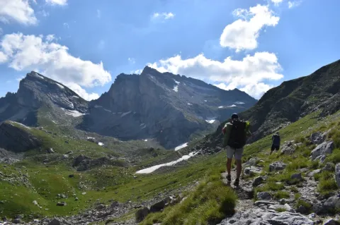 Ein Wanderer geht auf einem schmalen Pfad durch hügelige Landschaften. Einige Altschneefelder sind zu sehen. 