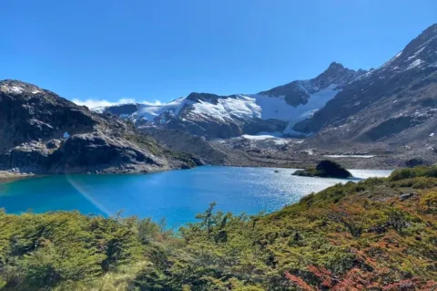 Blick auf einen blauen Bergsee. Vor dem See ist eine dunkelgrüne Wiese zu sehen, Im Hintergrund schneebedeckte Hügel. 