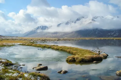 Ein hellblauer Fluss fließt auf einer flachen Ebene durch eine grüne Ebene. Im Hintergrund ist eine hügelige Landschaft zu sehen. Am Himmel sind Wolken zu sehen. 