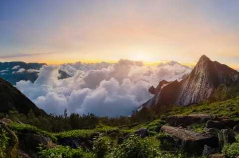 Blick auf einen steilen und felsigen Gipfel vor dem eine weiße Wolkendecke schwebt. Im Vordergrund sind grün bewachsene Felsen zu sehen. Am Horizont ist die Sonne bereits untergegangen, der Himmel ist orange und gelb gefärbt. 