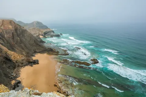 Weiter Blick auf eine Küstenlandschaft. Das türkise Meer schlägt an einen Sandstrand hinter dem eine steile Felswand steht. 