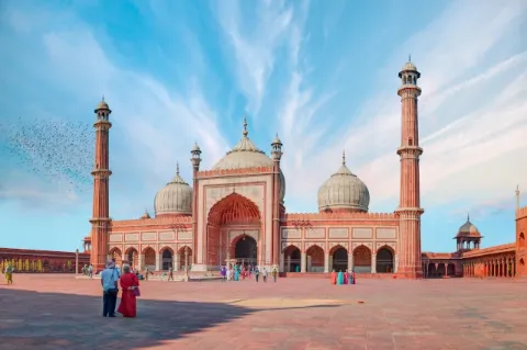 Es ist die Frontalansicht einer großen Moschee mit zwei hohen Türmen und runden Dächern zu sehen. Die verwendeten Steine sind rot. Vor der Moschee stehen zwei Menschen. 