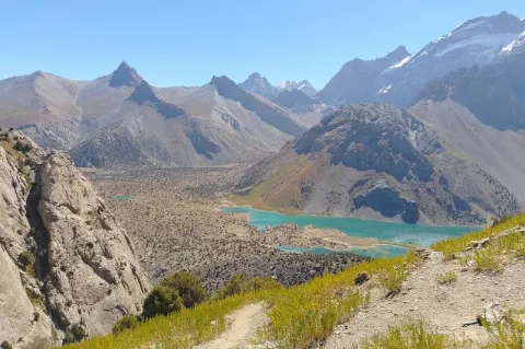 Aus der Distanz ist ein hellblauer, kleiner See zu sehen, der an eine felsige, karge Ebene angrenzt. Im Hintergrund sind spitz zulaufende Hügel zu sehen, die an manchen Stellen mit kleinen Waldstücken bewachsen sind. 