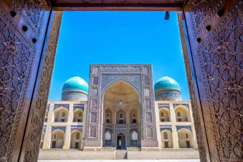 Blick durch ein massives, mit Ornamenten verziertes Tor auf eine Moschee mit blauem Mosaik. Links und rechts neben dem hohen Eingang sind zwei blaue Kuppeldächer zu sehen. 