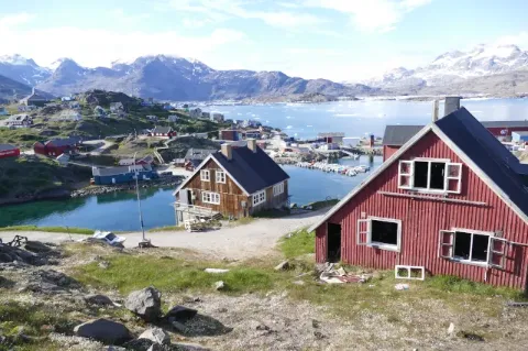Ein rotes Holzhaus steht auf einer teilweise mit Gras bewachsenen Fläche. Den Hang hinunter ist ein See zu sehen an dem ein braunes Holzhaus steht. Im Hintergrund sind mehrere steinige Hügel zu sehen. 