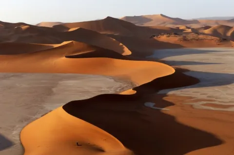 Blick auf eine geschwungene, steil zulaufende Düne, die in Kurven verläuft. Auf der rechten Seite der Düne wirft diese einen großen Schatten. Im Hintergrund sind weitere Sanddünen zu sehen.