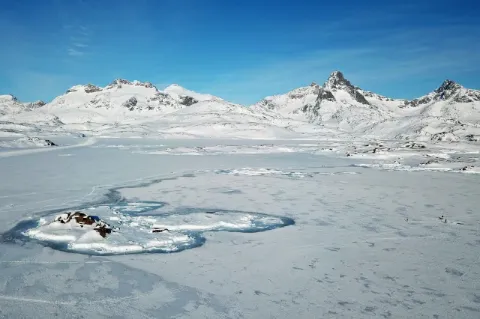 Blick über eine flache, große Eisfläche die mit Schnee bedeckt ist. Auf der Fläche ist eine kleine Siedlung zu sehen. Im Hintergrund erheben sich schneebedeckte, spitze Felsen.