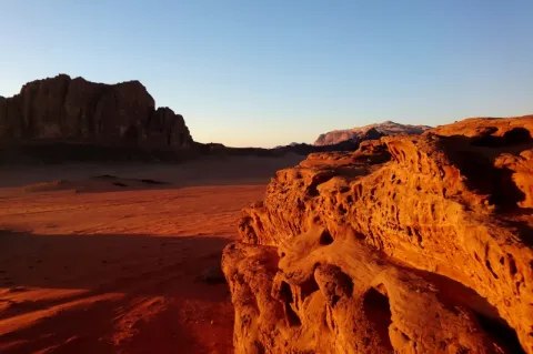 Blick über eine rote Sandstein und Sandlandschaft, die von der untergehenden Sonne angestrahlt wird und deshalb in goldgelbem Licht erstrahlt.