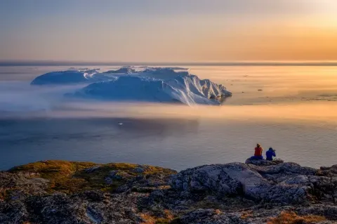 Zwei Menschen sitzen auf einem Felsen und schauen auf das Meer hinaus. Auf dem Meer ist der Teil eines Gletschers zu sehen und am Horizont geht die Sonne unter. 