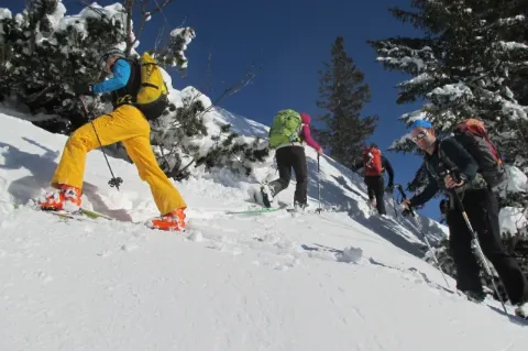 Mehrere Menschen in Winterausrüstung, Schneeschuhen und Skistöcken wandern einen steilen Schneehang hinauf. 
