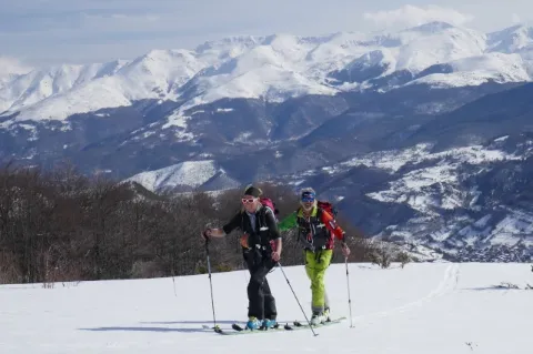 Zwei Skitourengeher gehen auf einer weißen Schneedecke und blicken in die Kamera. Im Hintergrund ist ein verschneites Gebirge zu sehen. 