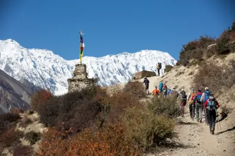 Auf der rechten Seite geht eine Wandergruppe auf einem steinigen Pfad auf einen kleinen buddhistischen Tempel zu. Im Hintergrund ist ein Teil des schneebedeckten Himalaya-Bergmassivs zu sehen. 