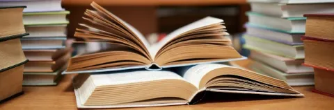 Zwei aufgeschlagene Bücher liegen auf einem Tisch und sind von weiteren Bücherstapeln umgeben