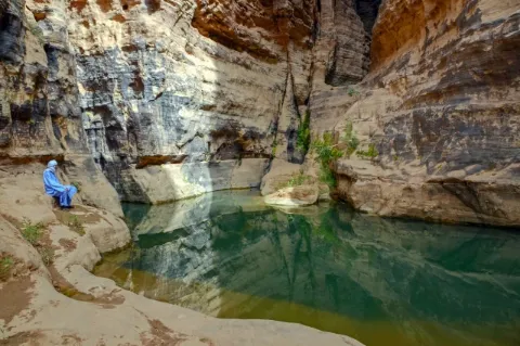 Ein Tuareg sitzt an einem kristallklarem Bergsee und schaut auf den See. Die Bergwände sind farbig. 