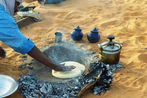 Ein Tuareg bereitet Fladenbrot mit seiner Hand in einer heißen Grube im Sand, gefüllt mit Holzkohle, zu. 