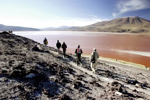 Wanderer laufen entlang eines steinigen Pfades und schauen auf das rot scheinende Wasser. 