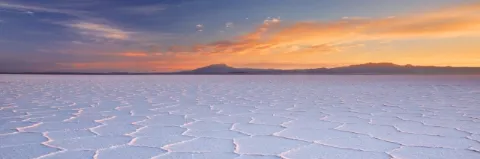 Der Himmel ist vom Sonnenuntergang rot gefärbt, darunter sind Berge. Davor ist ein Salzsee. 