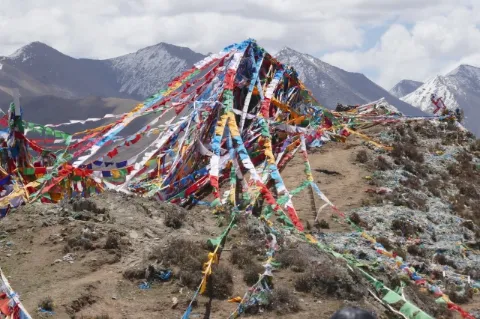Auf dem Gipfel eines braunen Berges sind bunte Gebetsfahnen. Dahinter sind schneebedeckte Berge.