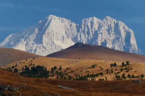 Ein großer, grau-weißer Berg am Horizont. Davor sind kleinere orange-braune Berge mit Bäumen.
