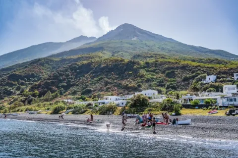 Menschen baden am Strand. Im Hintergrund sind ein paar Häuser und weit hinten ein Vulkan.