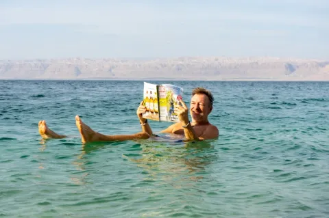 Ein Kunde liegt im Wasser und liest sich eine "Bike Bild" Zeitschrift durch. Im Hintergrund sind Berge. 