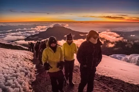 Mehrere dick eingepackte Wanderer steigen auf einen Berg hinauf. Die Sonne geht gerade auf.