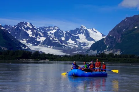 Fünf Menschen sitzen in einem Schlauchboot im Wasser. Im Hintergrund sind schneebedeckte Berge.