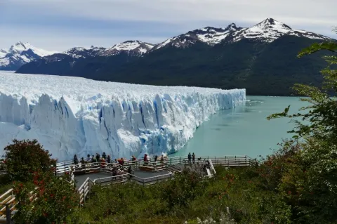 Ein mächtiger, bläulicher Gletscher ragt ins Wasser. Personen betrachten ihn von einer Plattform.
