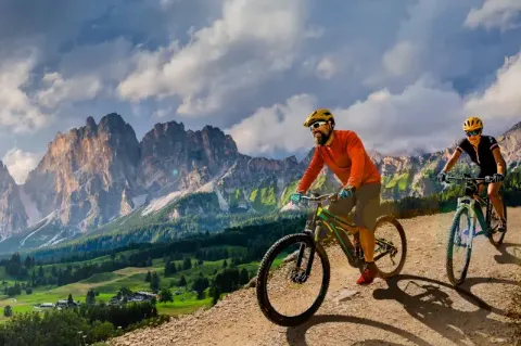Ein Paar radelt auf E-Bikes auf einer Schotterpiste vor grünen Wiesen und einer Bergkulisse im Hintergrund bei bewölktem Himmel