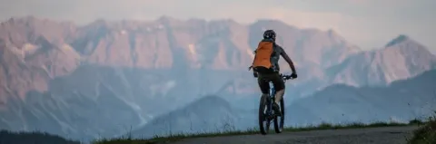 Mountainbiker mit Rucksack fährt über eine Teerstraße mit Blick auf die Bergkette bei leicht trübem Wetter.