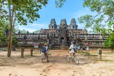Blick auf einen Tempel vor dem zwei Radfahrer auf sandigem Boden stehen.