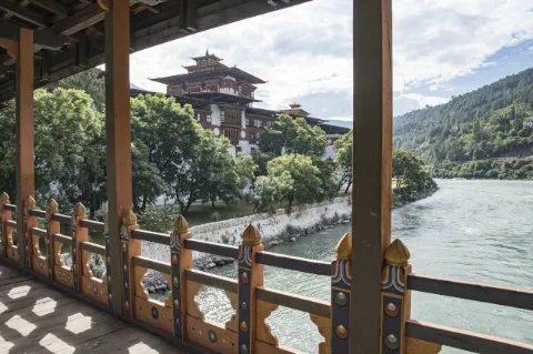 Blick von einer Brücke aus über einen Fluss auf ein buddhistisches Kloster. Vor dem Dzong wachsen grüne Bäume.