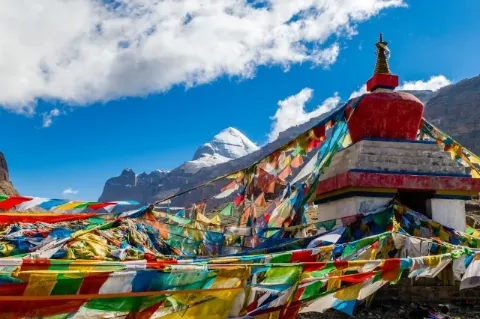 Blick auf einen schneebedeckten, spitzen Berggipfel im Hintergrund. Im Vordergrund sind Teile eines buddhistischen Tempels zu sehen, der mit vielen bunten Gebetsflaggen geschmückt ist. 