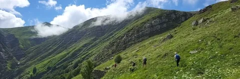 Zwei Wanderer laufen schräg an einem grünen Berghang entlang unter leicht bewölktem, blauen Himmel