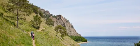 Zwei Wanderer laufen hintereinander auf einem Pfad zwischen grüner Wiese und blauem See
