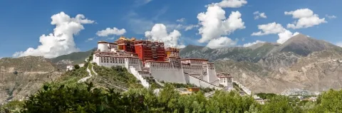 Auf einem Hügel steht ein weiß-roter Palast. Es gibt viele Bäume. In der Ferne sind hohe Berge.