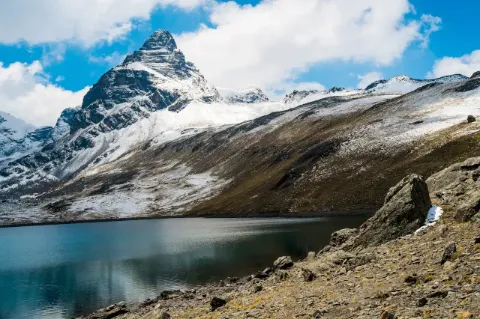 Blick über einen blauen Gebirgssee zum spitzen und felsigen Gipfel des Alpamayo, der an vielen Stellen noch mit etwas Schnee bedeckt ist.
