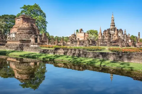 Blick vom Wasser auf einen historischen Park mit kleinen Gebäuden. Um den Park herum wächst viel Grünes.