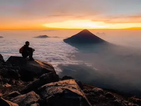 Man sieht links einen Wanderer auf einem Stein auf einem Berg über den Wolken mit orange-gelben Himmel im Hintergrund sitzen.