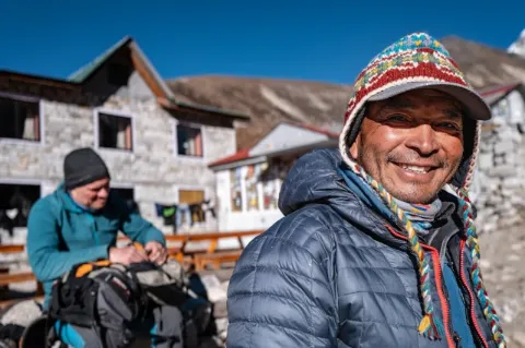 Der nepalesische Reiseleiter Harka Tamang blickt lächelnd in die Kamera. Er hat eine blaue Daunenjacke und eine Mütze mit bunten Mustern an und steht vor einem grauen Gebäude. 