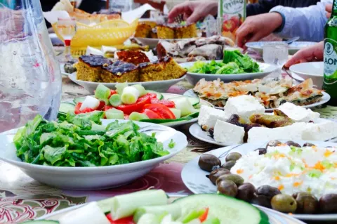 Verschiedenes frisches Essen aus dem Balkan steht auf einem Tisch. Um den Tisch sitzen Leute.