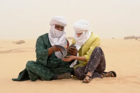 Zwei Menschen mit Kopftüchern sitzen im Sand und schauen sich gemeinsam ein Magazin an.