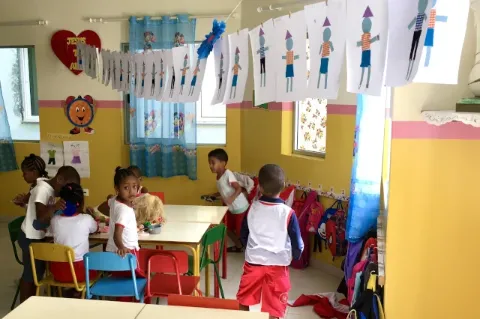 Ein Tisch steht in einem Raum. Kinder sitzen um den Tisch und malen, andere Kinder laufen um den Tisch.